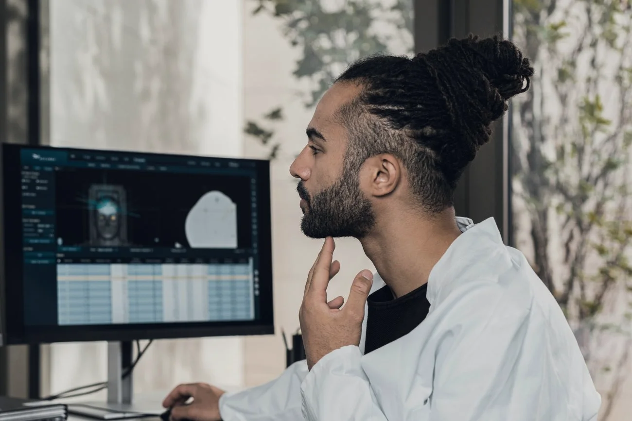 A specialist in a white coat studies scan images and a data table on a monitor.