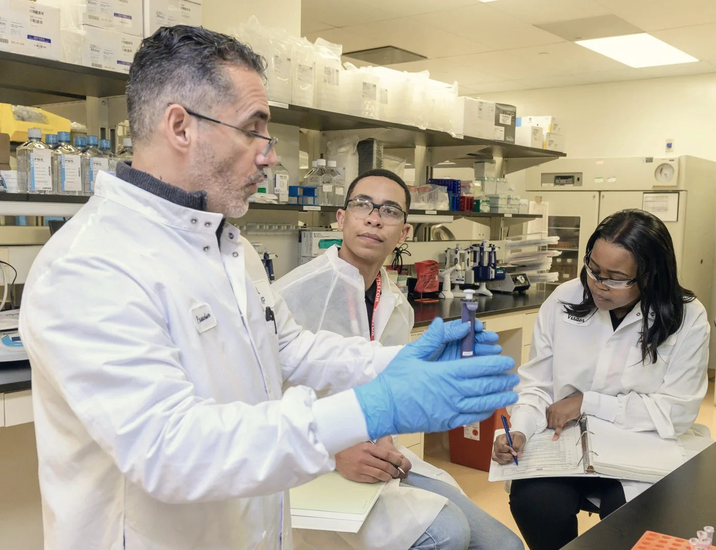 A scientist demonstrates a pipette to two colleagues taking notes in a laboratory.