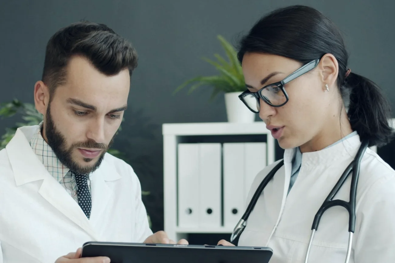 Two clinicians in white coats discuss information on a tablet.