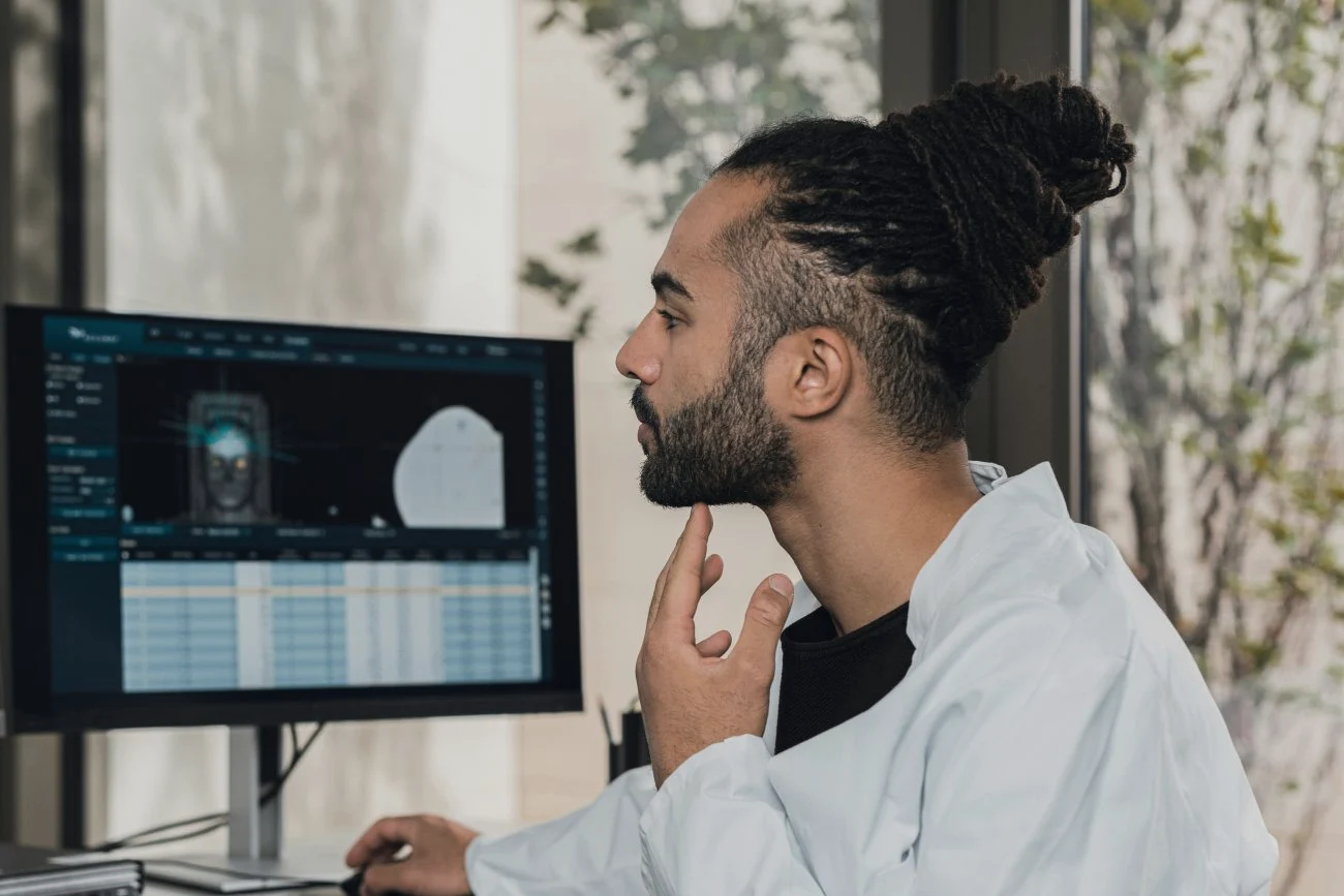 A specialist in a white coat studies scan images and a data table on a monitor.