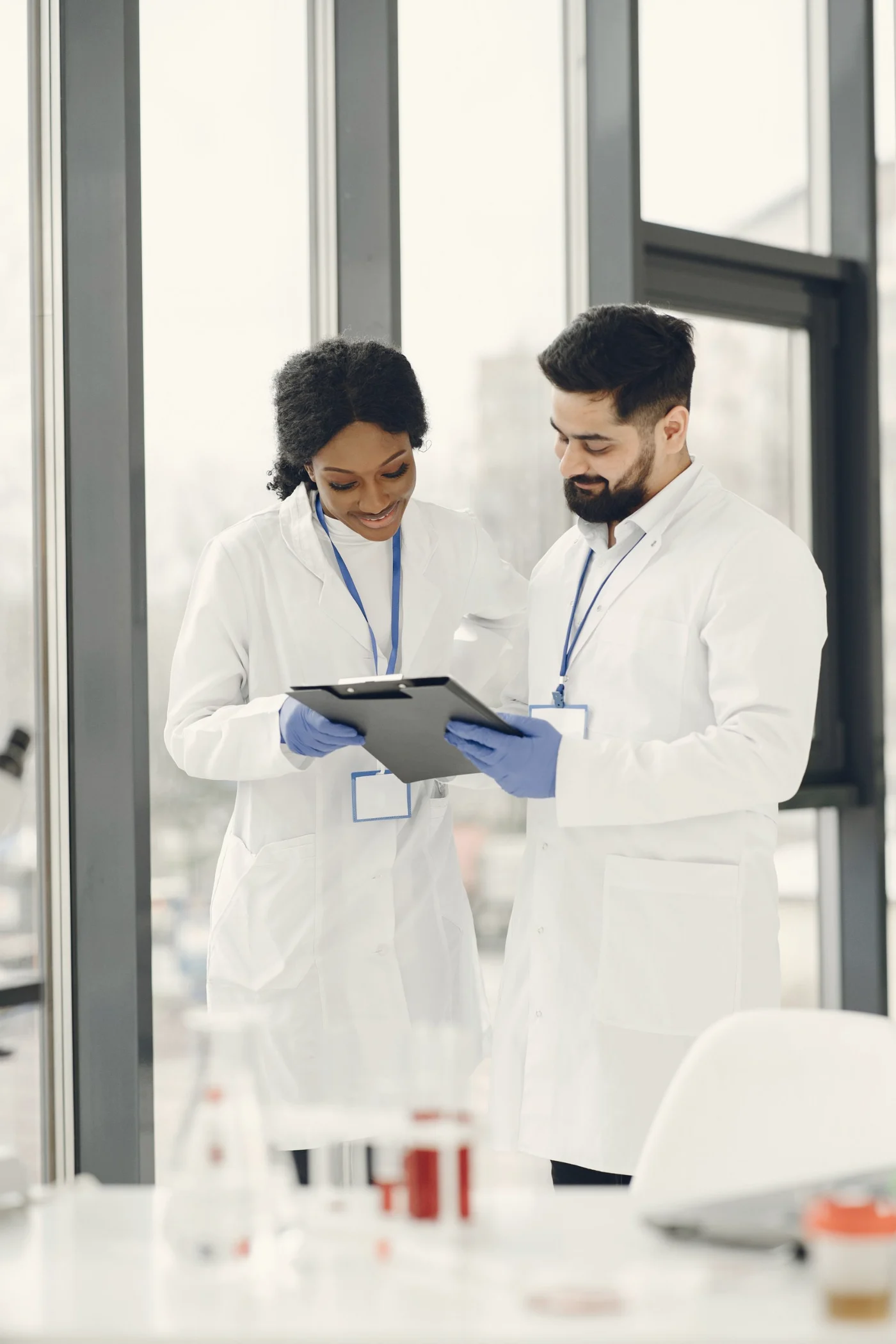 Two scientists in lab coats review notes on a clipboard by a window.